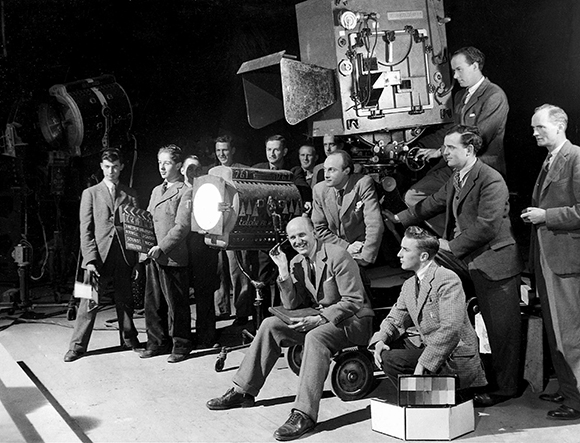 A black and white group portrait of men in suits surrounding large film cameras and studio lights.