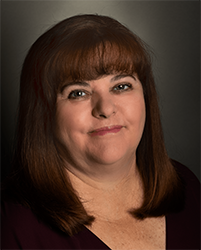 A woman with shoulder-length brown hair and bangs looks toward the camera against a dark background.