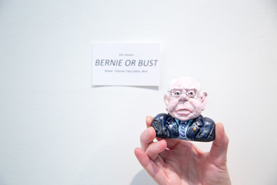 A hand holds a small, caricature clay bust of a man in a suit against a white wall with a label.