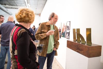 People standing in a white gallery space looking at framed photographs and a brass sculpture on a pedestal.