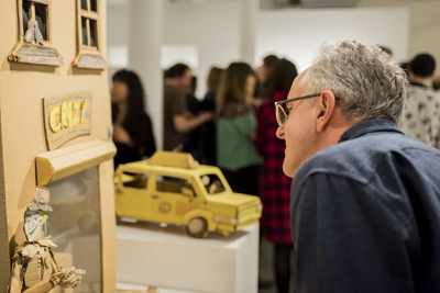 A man with glasses observes a miniature cardboard house and a yellow toy van in a gallery setting.