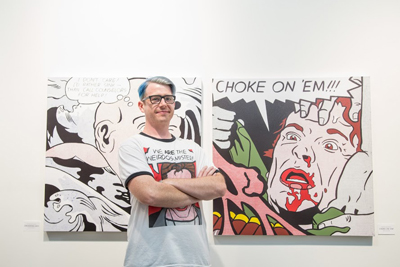 A man with blue hair and glasses stands with arms crossed in front of two large comic-style art pieces.
