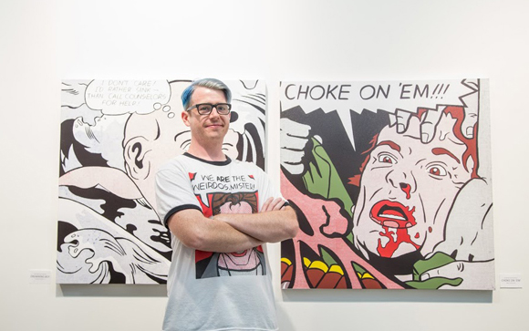 A man with blue hair and glasses stands with arms crossed in front of two large comic book style paintings.