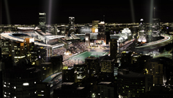 Aerial night view of a large, illuminated stadium surrounded by city skyscrapers and bright spotlights.