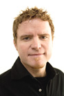 A man with short, wavy reddish-brown hair and a small goatee wearing a black collared shirt.