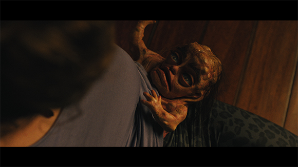 A small, pale, distorted creature with large eyes clings to the back of a person wearing a purple shirt.