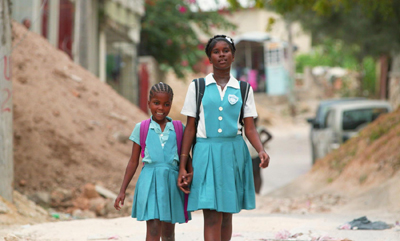 Two girls in teal school uniforms walk together down a dusty street in a residential area.