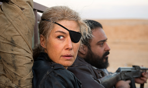 A woman with an eye patch and a man with a beard sit in a vehicle in a desert landscape.