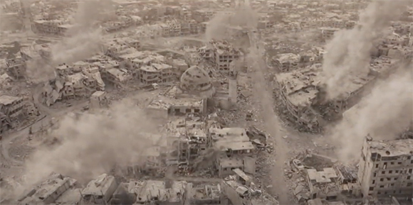 Aerial view of a city with heavily damaged buildings and plumes of smoke rising from the rubble.