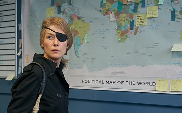 A woman with an eye patch looks to the side in front of a large political map of the world.