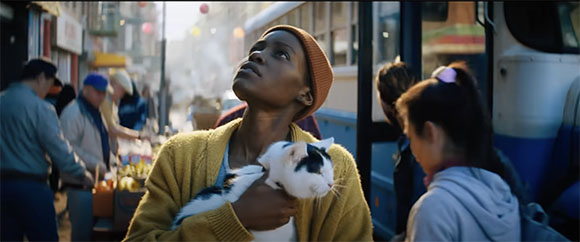 A woman in a yellow cardigan holds a black and white cat while looking up in a busy outdoor market.