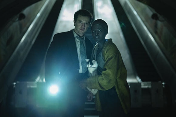 A man in a suit and a woman in a yellow robe hold a small cat in a dark, metallic tunnel.
