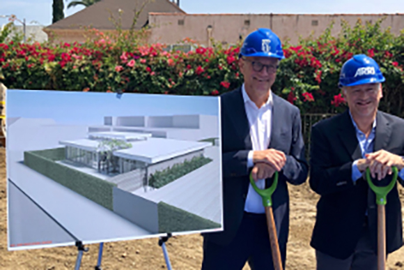 Two men in suits and blue hard hats hold shovels next to a large architectural rendering of a modern building.