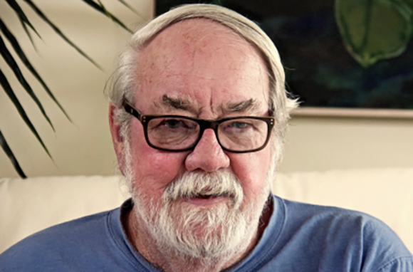 A man with white hair, a white beard, and black-rimmed glasses wearing a blue shirt.