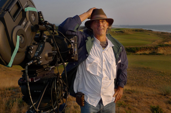 A man in a brown hat and blue jacket stands beside a large professional film camera on a grassy cliffside.