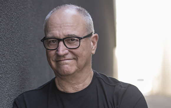 A man with short graying hair and black-rimmed glasses smiling slightly against a dark wall.