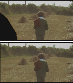 A person in a green jacket and brown bag stands in a field facing haystacks.
