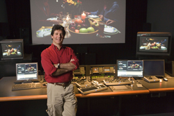A man in a red shirt and khaki pants stands with arms crossed in a dark video editing suite.