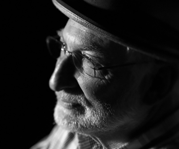 Black and white profile portrait of an elderly man wearing glasses and a brimmed hat in dramatic lighting.
