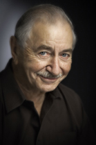 A close-up portrait of an elderly man with graying hair and a mustache, smiling slightly against a dark background.