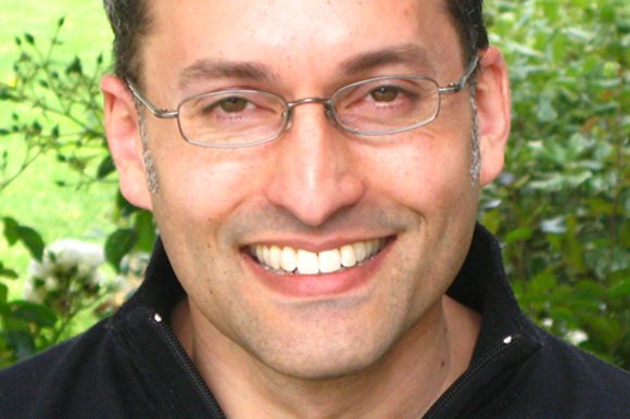 A man with short dark hair and glasses smiling in front of green foliage.