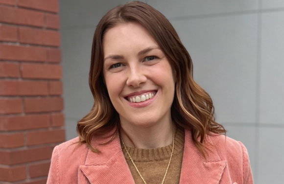 A smiling woman with wavy brown hair wearing a pink corduroy blazer and a brown sweater.