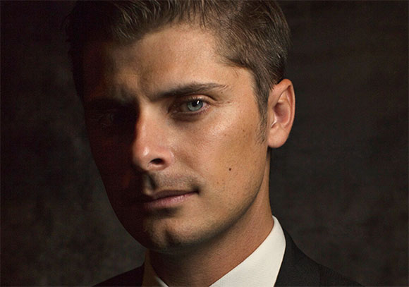 Close-up portrait of a man with light eyes and short brown hair looking toward the camera.
