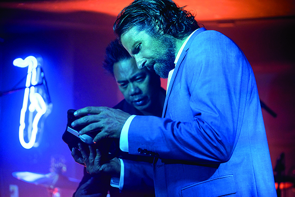 A man in a light suit plays a handheld electronic instrument under blue and red stage lights.