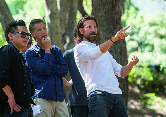 A man in a white shirt points toward something while two other men look on in a wooded setting.