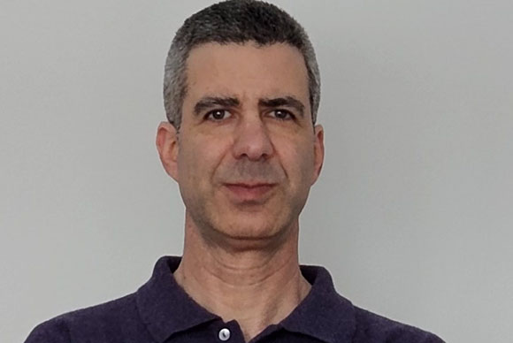 A man with short salt-and-pepper hair and dark eyes wearing a purple polo shirt against a plain background.