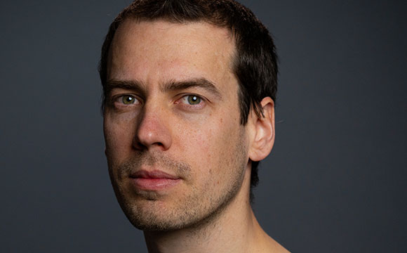 Close-up portrait of a man with short dark hair and light eyes looking forward against a dark background.