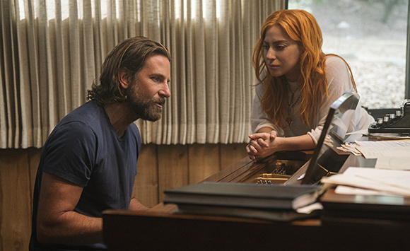 A man in a blue shirt and a woman with red hair sit at a wooden piano in a room with curtains.