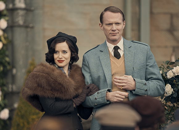 A woman in a black hat and fur stole stands beside a man in a light blue blazer and tan sweater vest.