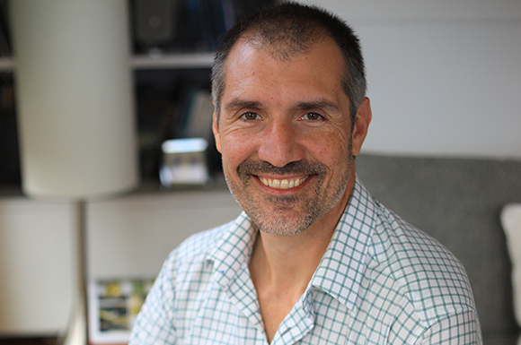 A smiling man with short dark hair wearing a white and blue checkered button-down shirt.