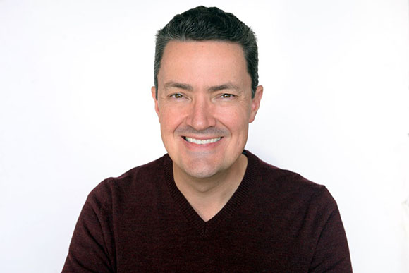 A smiling man with short dark hair wearing a maroon V-neck sweater against a white background.