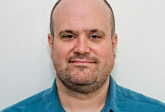 A man with short hair and a light beard smiles slightly against a plain white background.