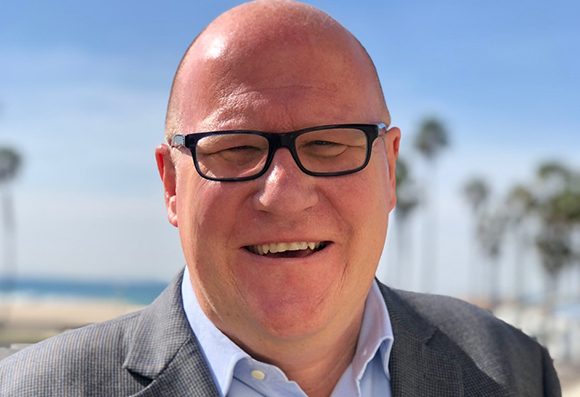 A smiling bald man with black-rimmed glasses wearing a grey suit jacket and light blue shirt.
