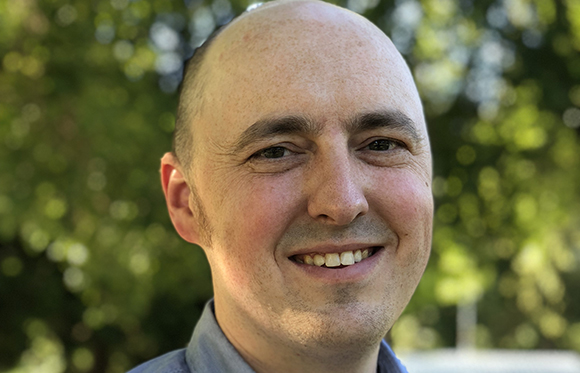 A smiling bald man with light skin and a blue shirt against a blurred green background.