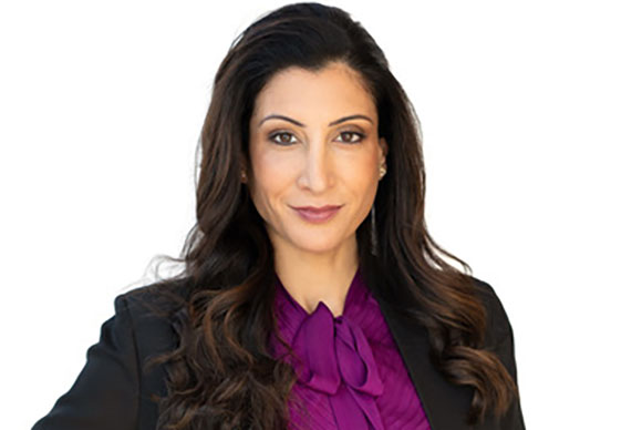 A woman with long, wavy dark hair wearing a purple blouse and a black blazer against a white background.