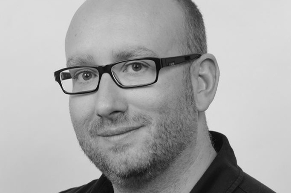 Black and white close-up portrait of a man wearing dark-rimmed glasses.
