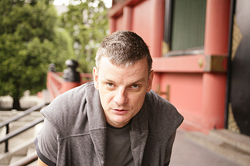 A man with short hair wearing a grey vest looks directly into the camera.