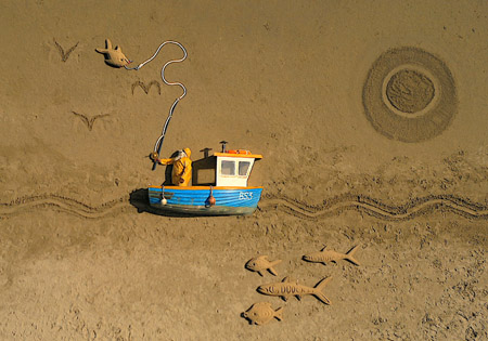 A blue toy boat sits on sand next to sand drawings of fish and a large circle.