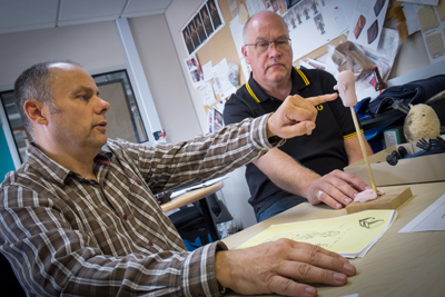 Two men sit at a desk, one pointing at a small pink model mounted on a wooden stick.
