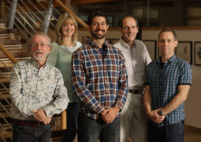 Five smiling adults standing in a group indoors near a wooden staircase.
