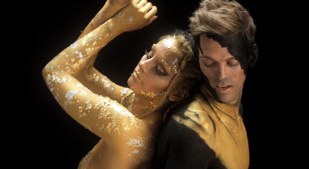 A woman in gold body paint leans against a man with black and gold body paint.