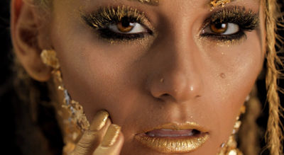 Close-up of a person with gold glitter eyeshadow, gold lipstick, and gold leaf accents on their skin.