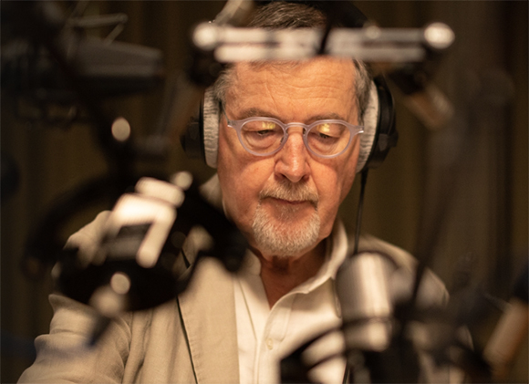 A man with glasses and headphones looks down while surrounded by studio microphones.