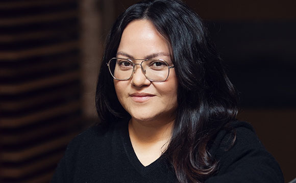 A woman with long dark hair and wire-rimmed glasses smiling slightly against a dark, blurred background.