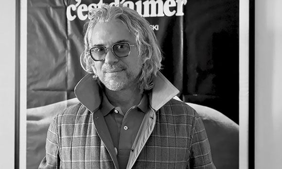 Black and white portrait of a man with wavy hair and glasses wearing a collared shirt and quilted vest.