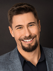 A smiling man with short brown hair and a dark goatee wearing a grey blazer and black shirt.